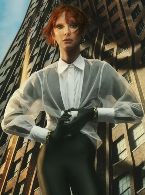 Stylish woman with red hair wearing a white shirt, gloves, and bracelet, posing against a building in an urban fashion portrait.