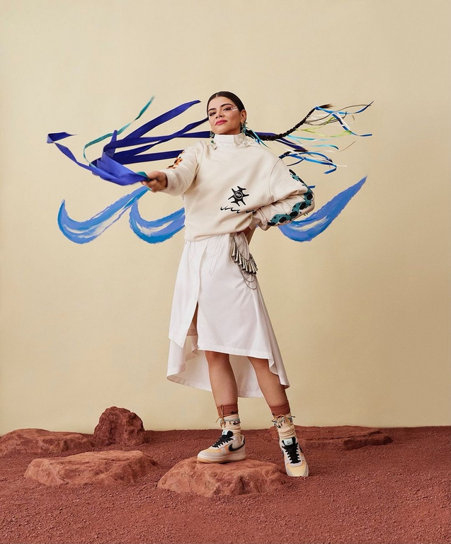 Young woman posing in a cream-colored sweatshirt and white skirt, adorned with blue ribbons in her hair, standing on red rocks.