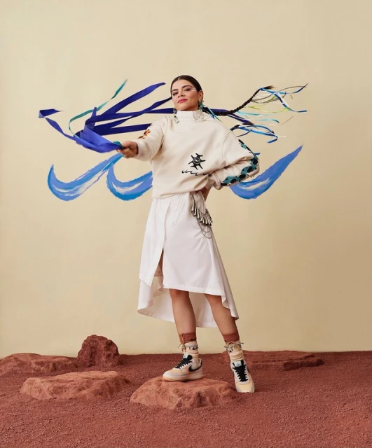 Young woman posing in a cream-colored sweatshirt and white skirt, adorned with blue ribbons in her hair, standing on red rocks.