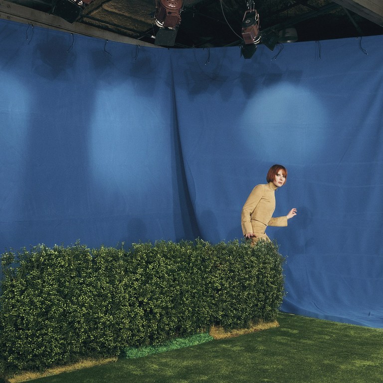 Woman in tan outfit emerges from behind a green hedge in a photo studio with artificial grass and blue backdrop.