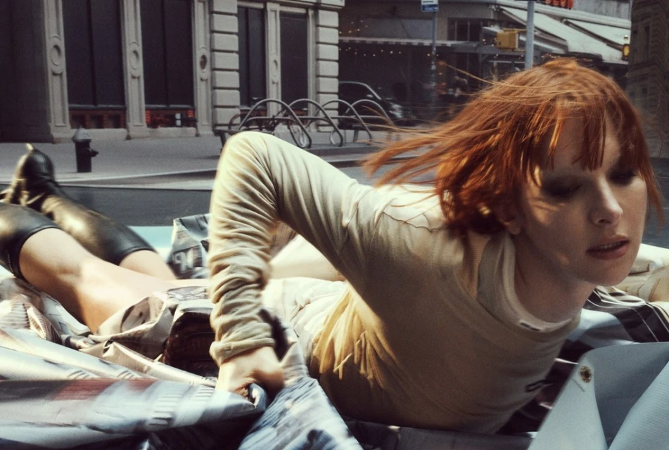 Adult female with red hair and black boots laying in a pile of clothes in a city street with buildings in the background.