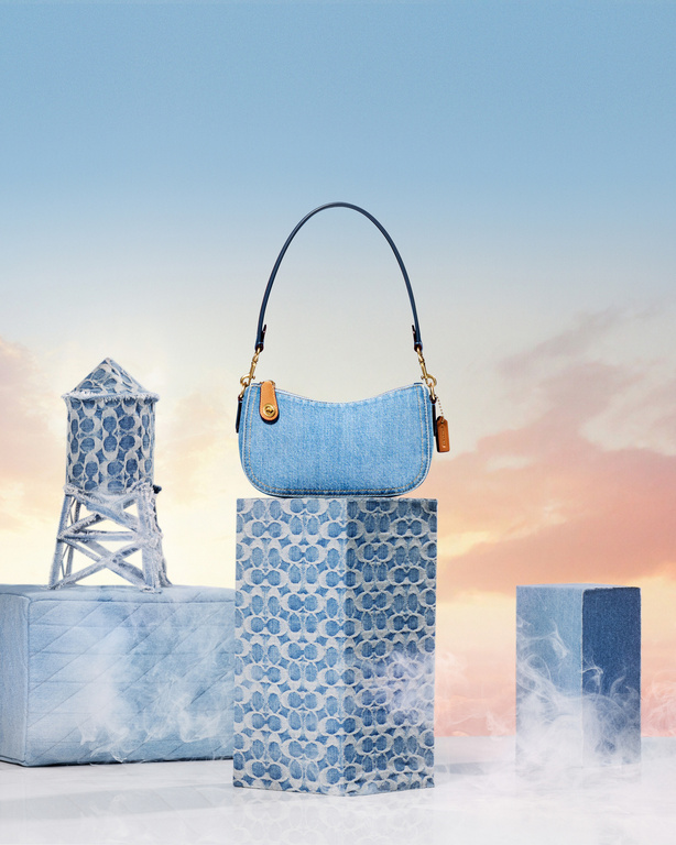 Denim Coach purse sitting on a display block with a denim water tower and soft blue and pink sky in the background in a studio setting.