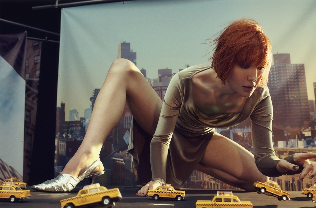 Woman in dress posing with miniature yellow taxi cars in a staged photo shoot with a cityscape backdrop, creating a surreal scene.
