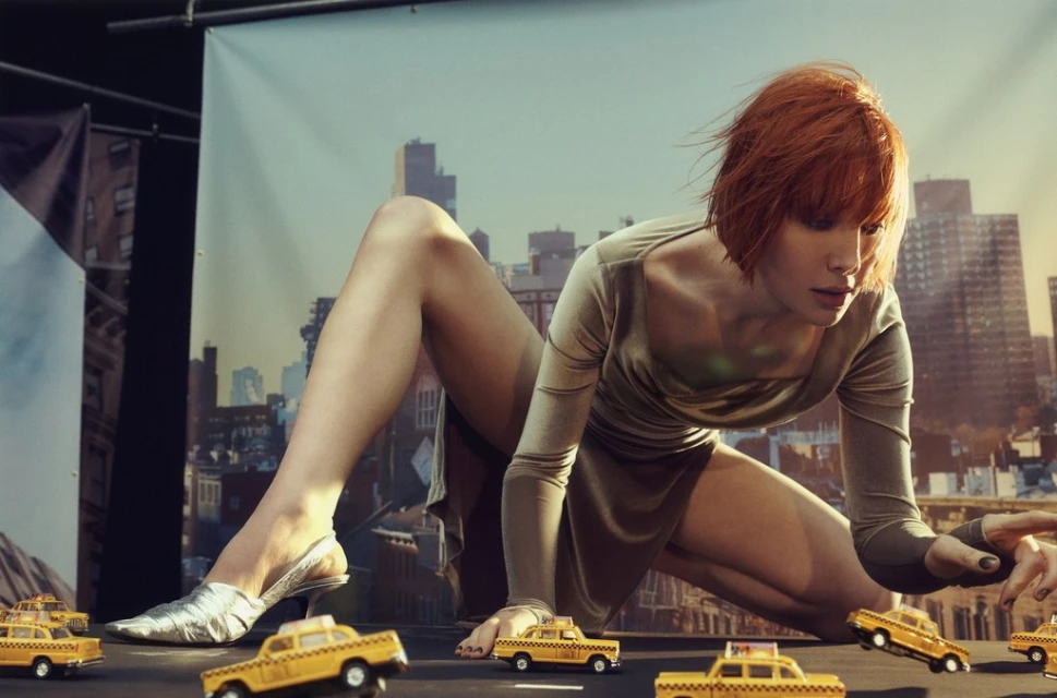 Woman in dress posing with miniature yellow taxi cars in a staged photo shoot with a cityscape backdrop, creating a surreal scene.