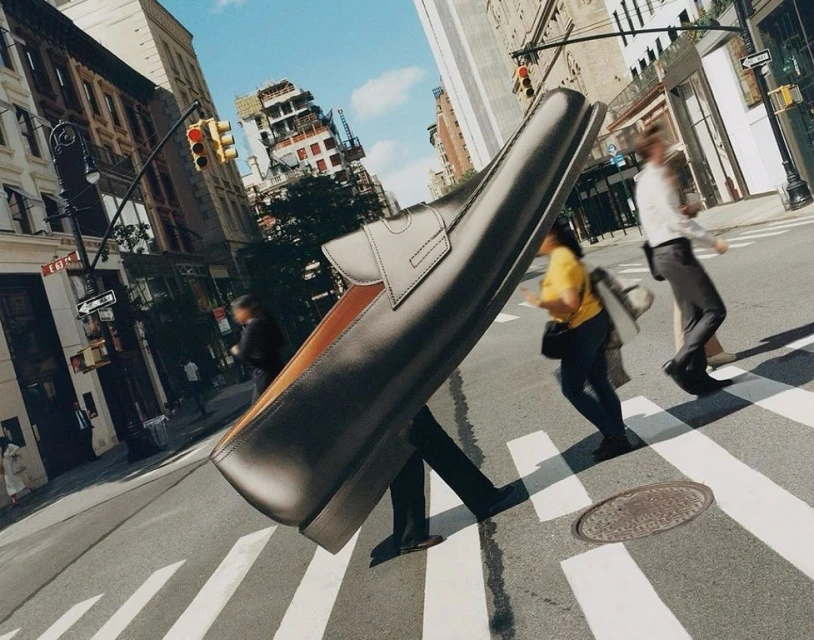 A giant loafer shoe stands in a pedestrian crosswalk with blurred pedestrians and buildings in background under a clear sky.