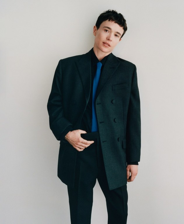 Elliot Page wearing a dark green oversized blazer, black shirt, and bright blue tie against a plain white background