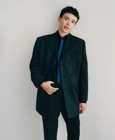 Elliot Page wearing a dark green oversized blazer, black shirt, and bright blue tie against a plain white background