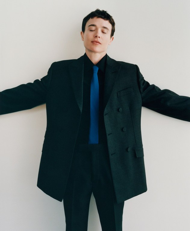 Adult male in a dark suit with a blue tie and closed eyes, stands against a plain white background with his arms spread.