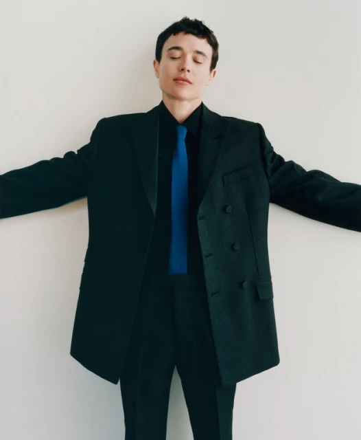 Adult male in a dark suit with a blue tie and closed eyes, stands against a plain white background with his arms spread.
