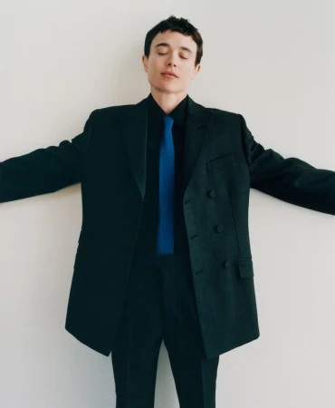 Adult male in a dark suit with a blue tie and closed eyes, stands against a plain white background with his arms spread.