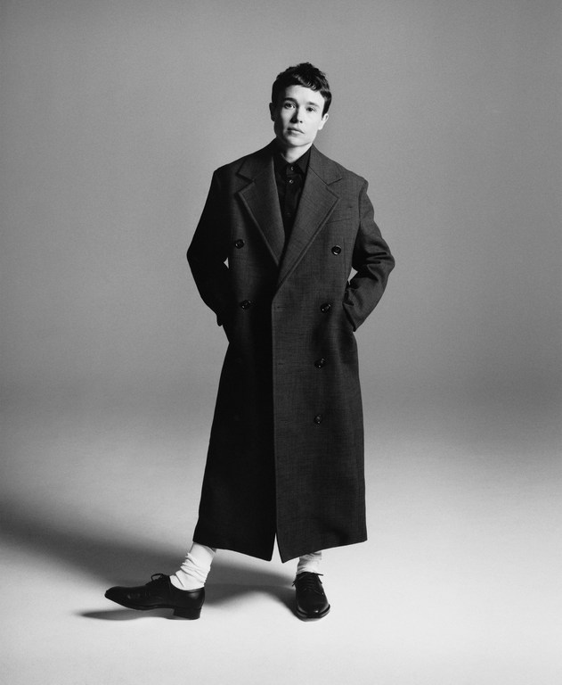 Elliot Page wearing a dark double-breasted trench coat, white socks, and black shoes in a studio portrait.
