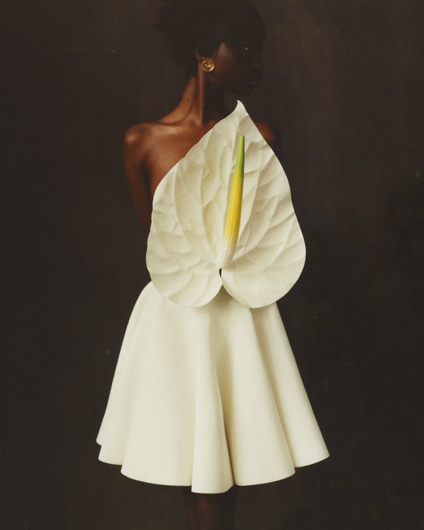 A dark-skinned woman in a white dress adorned with a large white Anthurium flower stands against a dark backdrop in a studio shot.