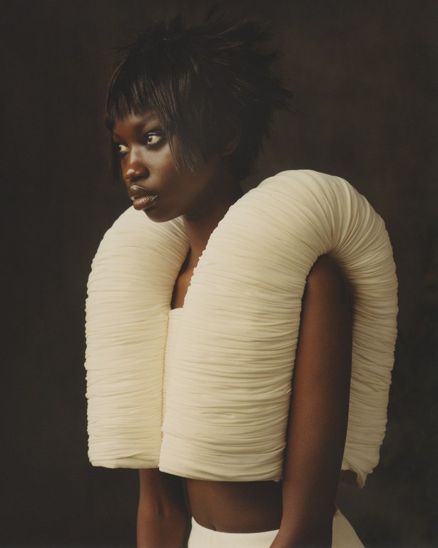 Portrait of a Black woman in a white architectural dress, standing in a studio with a dark background, looking to the left.