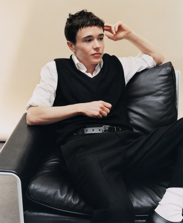 Portrait of Elliot Page sitting in a black leather chair, wearing a black sweater vest, white shirt, and black dress pants in a studio.