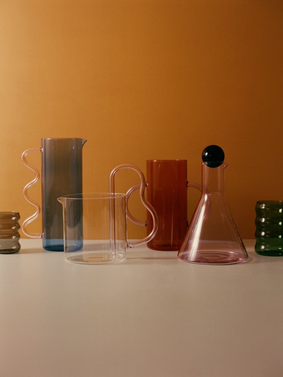 Still life shot of several colorful glassware pieces including pitchers and cups with rounded handles against a warm orange background.