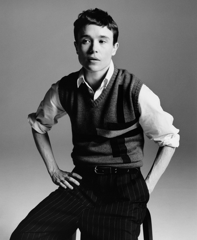 Black and white portrait of Elliot Page in a sweater vest and pinstripe pants, seated with hands on hips against a neutral backdrop.