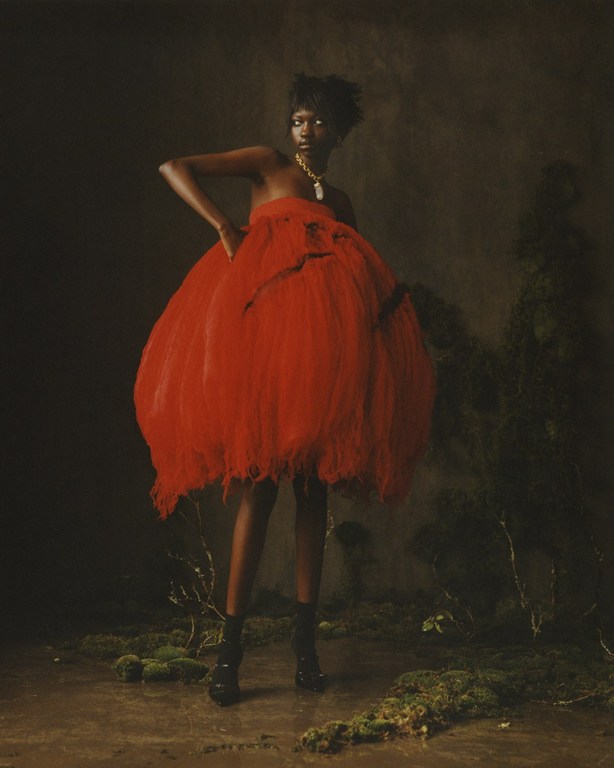 Model wearing a voluminous red dress and gold necklace posing in a moody, moss-covered set designed by Alicia Sciberras