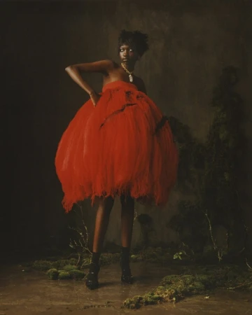 Model wearing a voluminous red dress and gold necklace posing in a moody, moss-covered set designed by Alicia Sciberras