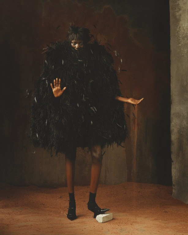 Fashion shot of a Black adult female in black feather dress standing indoors on red floor. A pillar appears on the right.