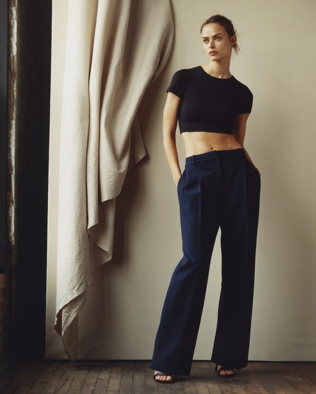 A slender woman poses in a black crop top and blue trousers in front of a neutral-toned wall with a draped fabric backdrop.