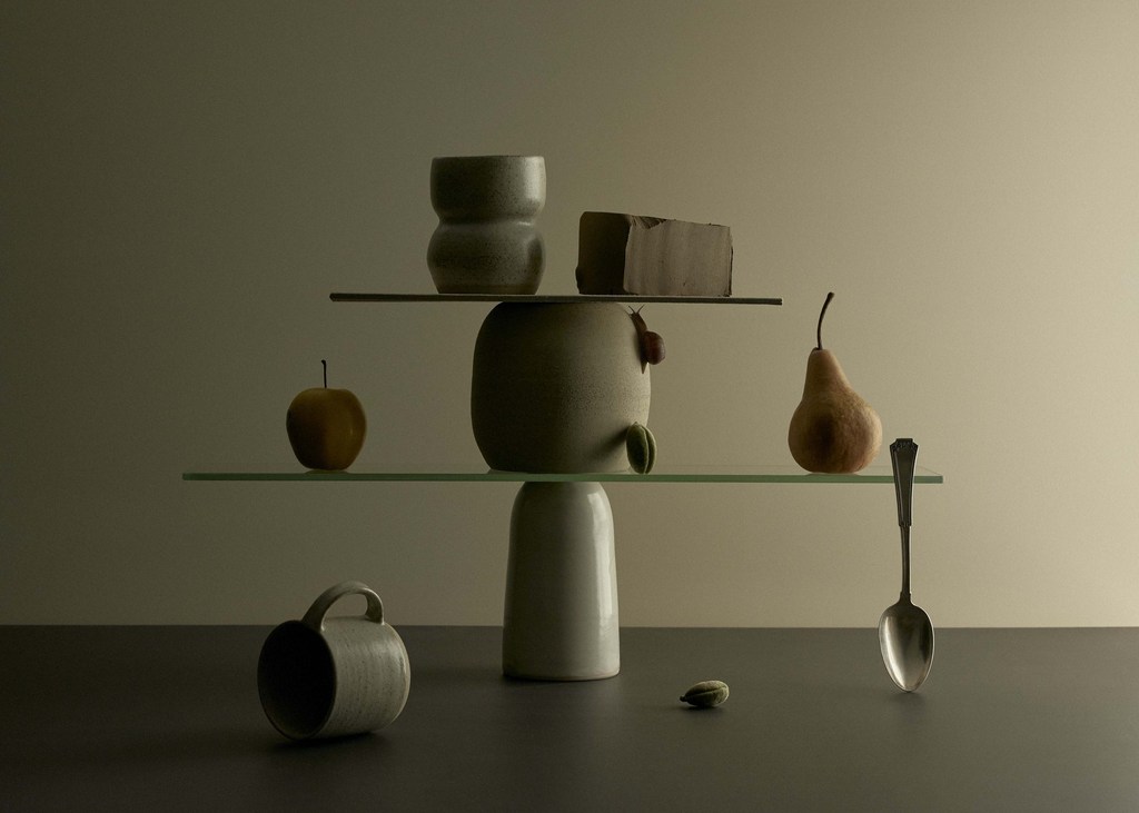 Still life photograph of balanced objects: vase, pear, apple, spoon, and chocolate bar, arranged on glass shelves in a minimalist composition.