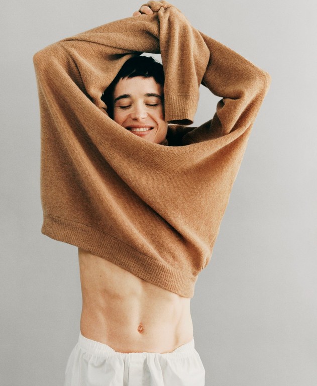 Adult male smiles while taking off a tan sweater indoors against a grey wall, showcasing his abs and wearing white shorts.