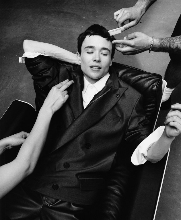 Black and white portrait of a non-binary adult lying in a chair while multiple sets of hands style their hair and adjust their clothing.