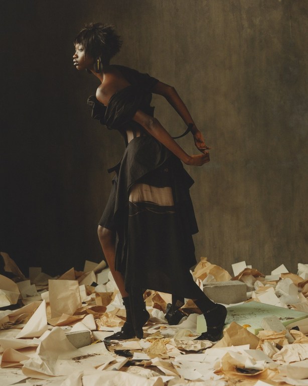 Fashion shot of a Black female model wearing an avant-garde black dress and booties, standing on crumpled paper in a studio setting.
