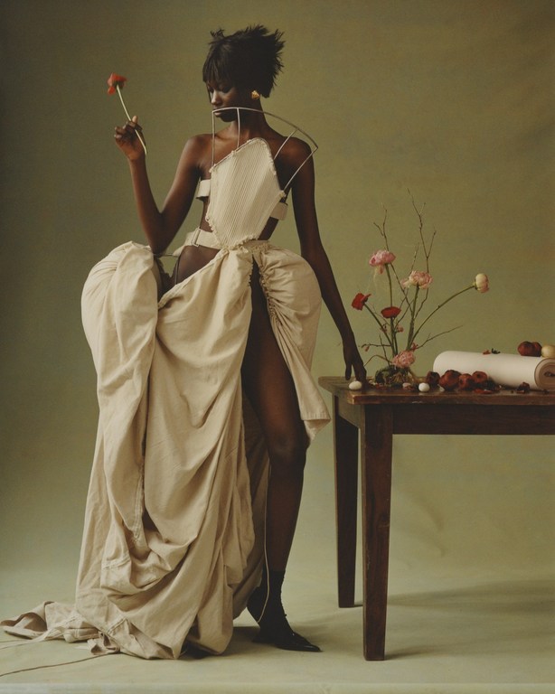 Elegant, dark-skinned woman in sculptural couture poses with a flower near a wooden table in a neutral-toned studio setting.