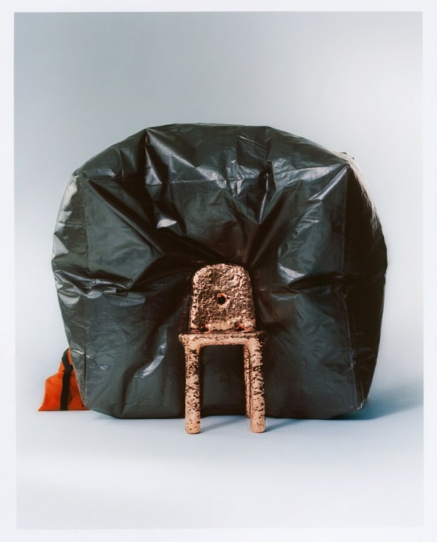Indoor studio shot of a child-sized chair crafted from textured bronze, positioned in front of a large black plastic bag.