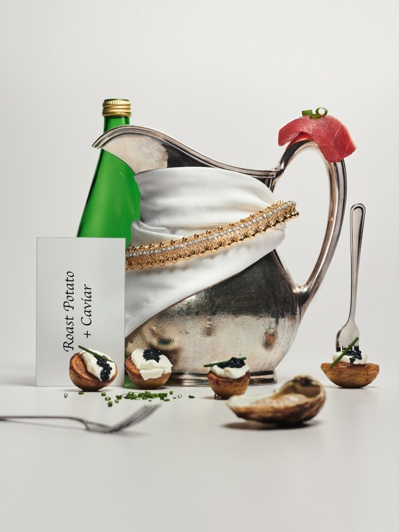Food still life of roast potato bites with caviar, silver gravy boat with fabric, and green bottle, set against a white background.