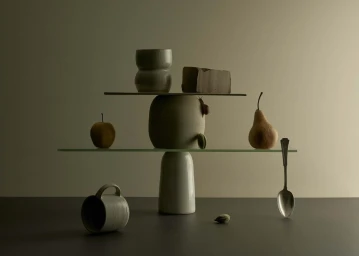 Balanced still life setup with ceramics, fruit, a snail, and a spoon on glass shelves against a muted background.