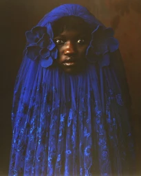 Portrait of a Black woman wearing a striking royal blue sheer hooded veil with large fabric flower appliqués by set designer Alicia Sciberras
