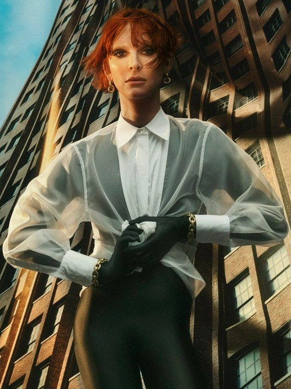 Low angle editorial portrait of a red-haired woman wearing a sheer white blouse, black gloves, and a tall building exterior background.