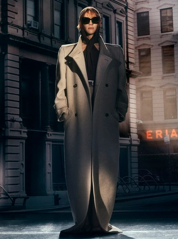 Model wearing dark sunglasses and a long beige structured coat standing on a city street.