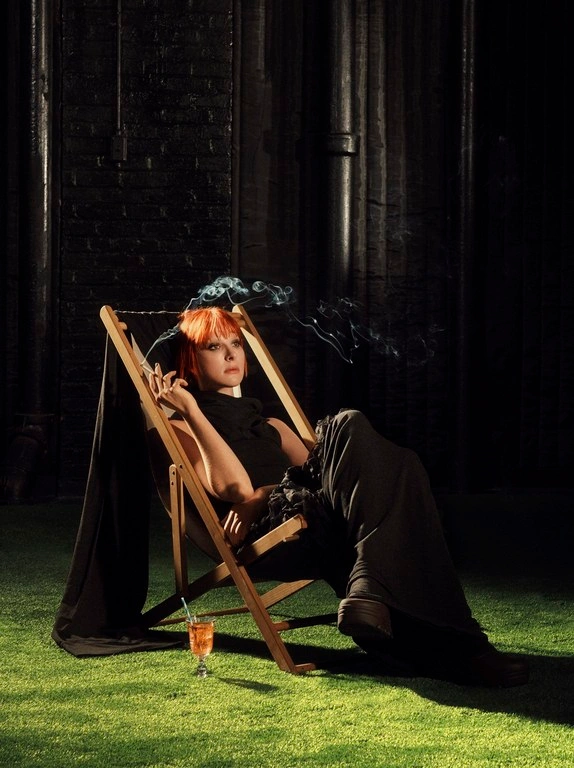Woman with red hair seated in a wooden deck chair holding a cigarette with smoke swirling against a dark brick wall.