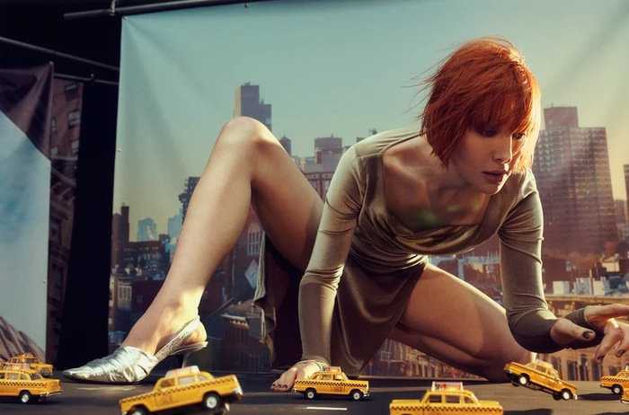 A red-haired woman crouching in a studio set with miniature toy taxis, designed by set designer Alicia Sciberras.