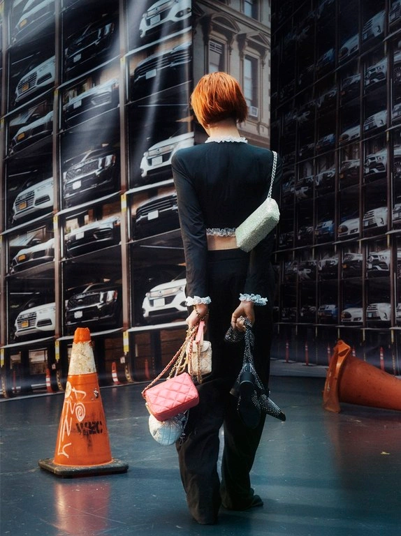 Woman with short reddish-orange hair wearing a black crop top and trousers holding multiple handbags standing before a parked car wall.