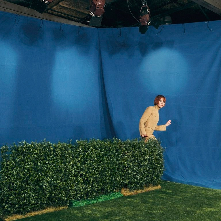Woman in beige top standing behind a low green hedge against a blue studio backdrop with overhead lights.