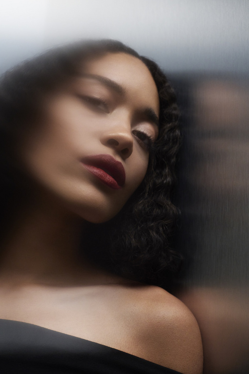 Close-up portrait of a woman with dark curly hair and red lipstick, wearing a black strapless top against a blurred grey background.
