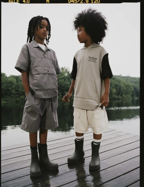 Two young boys standing on a wooden dock near a lake wearing casual outerwear and boots, portraying a relaxed outdoor style for kids.