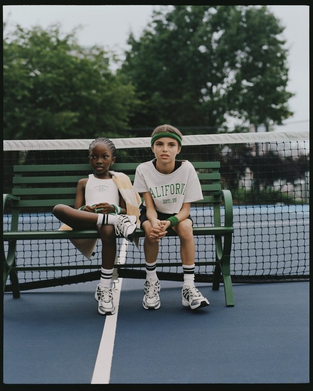 Two young girls, one Black and one White, sit on a green bench on a blue tennis court wearing sporty outfits and tennis shoes.