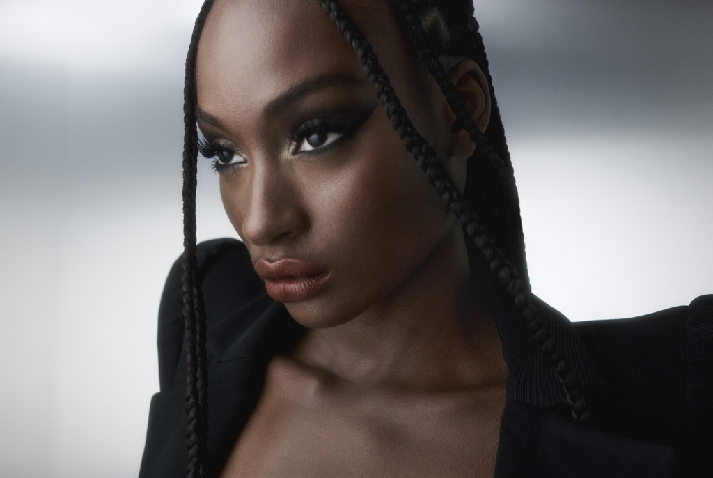 Close up portrait of a Black woman with braided hair wearing dark makeup and a black blazer against a grey backdrop