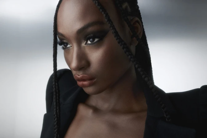 Close up portrait of a Black woman with braided hair wearing dark makeup and a black blazer against a grey backdrop