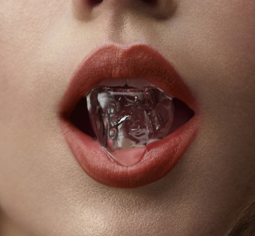 Close-up macro shot of lips wearing coral red lipstick holding a clear ice cube in the mouth by set designer Caz Slattery.