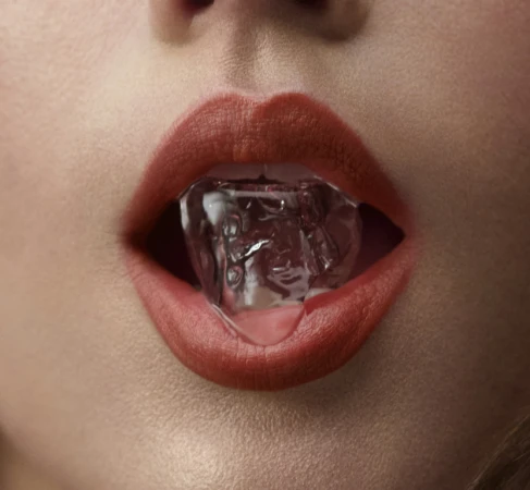 Close-up macro shot of lips wearing coral red lipstick holding a clear ice cube in the mouth by set designer Caz Slattery.