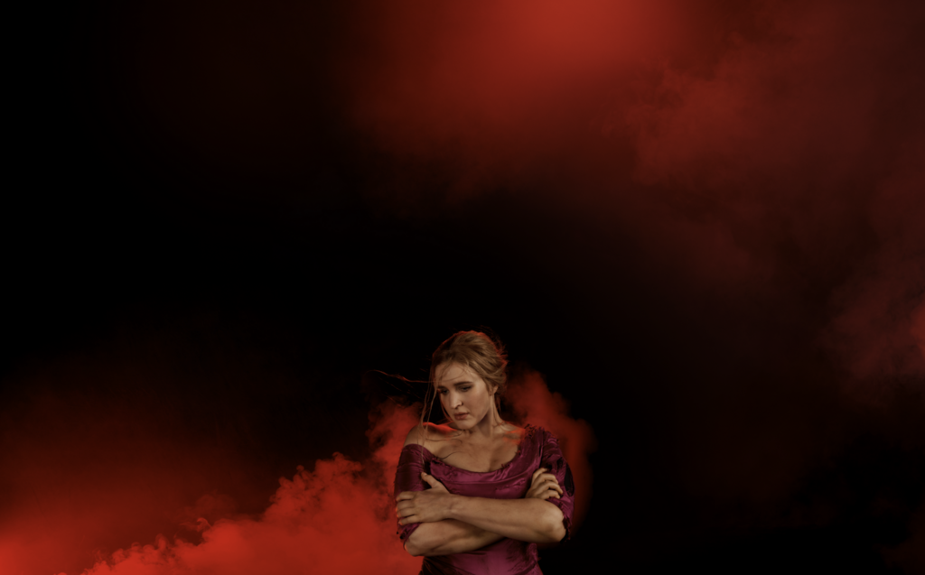Woman in a dark red dress embraces herself in a dramatic pose against a dark, smoky red background with creative lighting. 