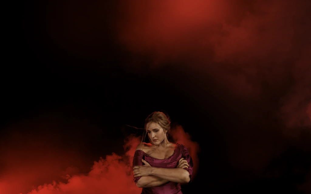 Woman in a dark red dress embraces herself in a dramatic pose against a dark, smoky red background with creative lighting. 