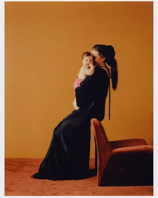 Woman in long black dress holds baby in pink, seated next to red velvet armchair on brown carpet against orange wall, studio lighting.