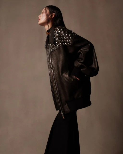 Studio shot of adult female model in an oversized studded leather jacket and black dress, posing and looking up against a neutral backdrop.